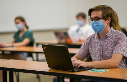 Photo of a student sitting at a desk with a face mask on looking at his laptop