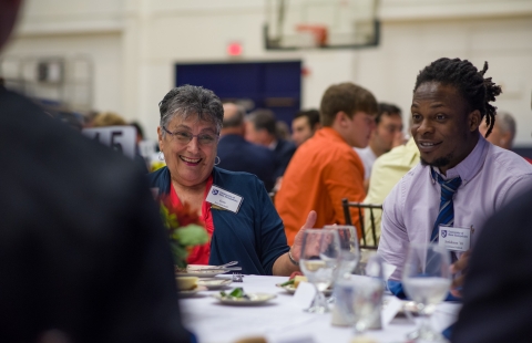 An elderly white woman and a young black man sitting at a dinner table smiling and laughing