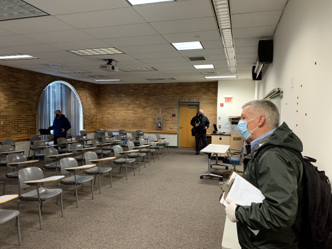 A man with gray hair and a blue mask and parka on stands with a clipboard in his hand looking at an empty classroom.